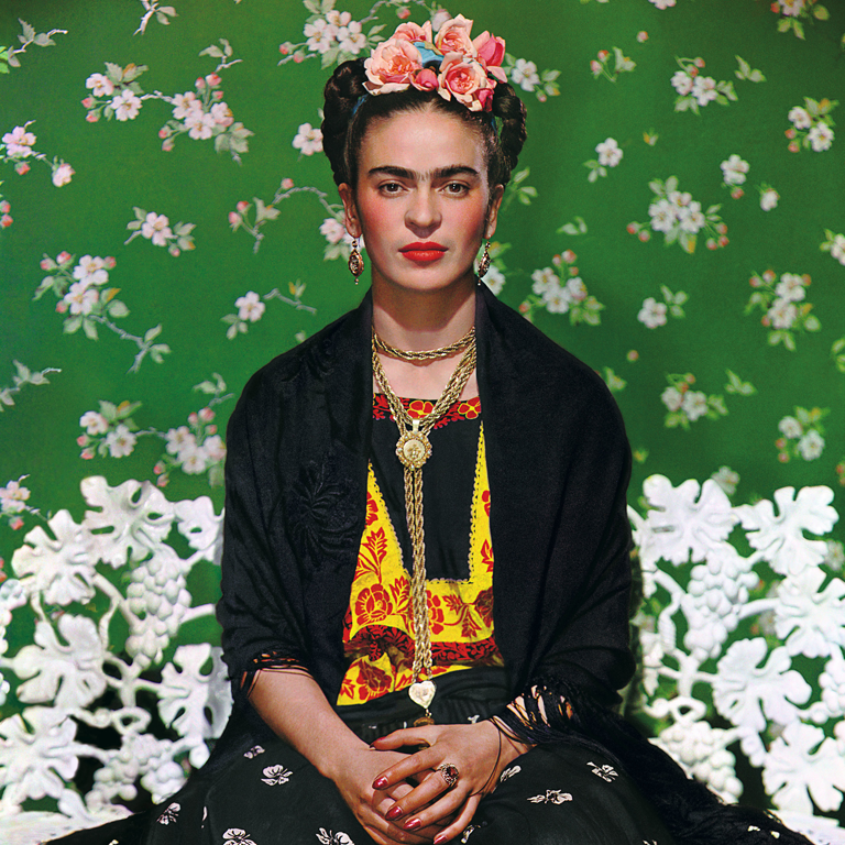 Becoming Frida Kahlo Photo of Frida Kahlo sitting on a fancy ornate chair in front of a wall of featuring pink flowers on green wallpaper. From the documentary Becoming Frida Kahlo.