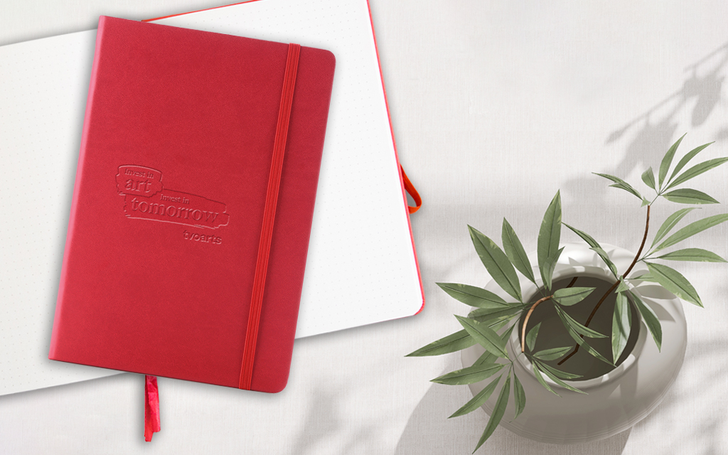 Photo of a closed red notebook sitting on top of an open red notebook with a plant in a vase sitting beside it.